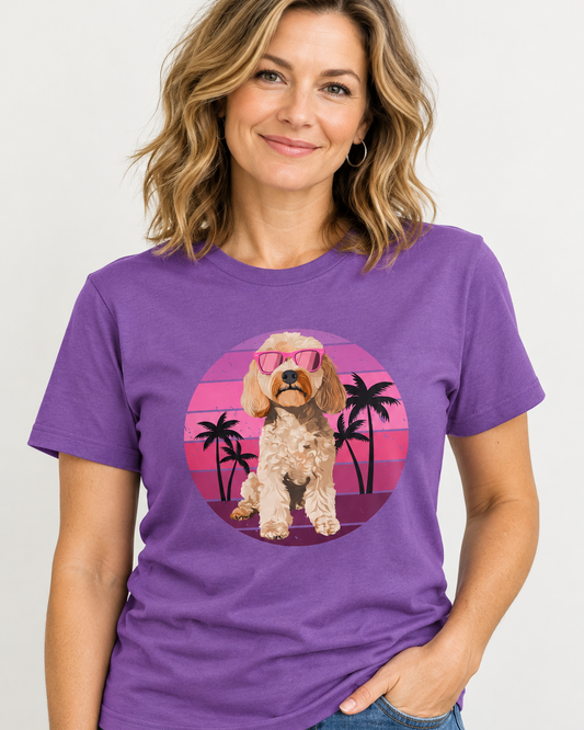 A woman models a royal purple short-sleeve crewneck t-shirt featuring a circular graphic of a fluffy cream-coloured cockapoo dog wearing pink sunglasses, set against a pink and purple sunset with palm trees.