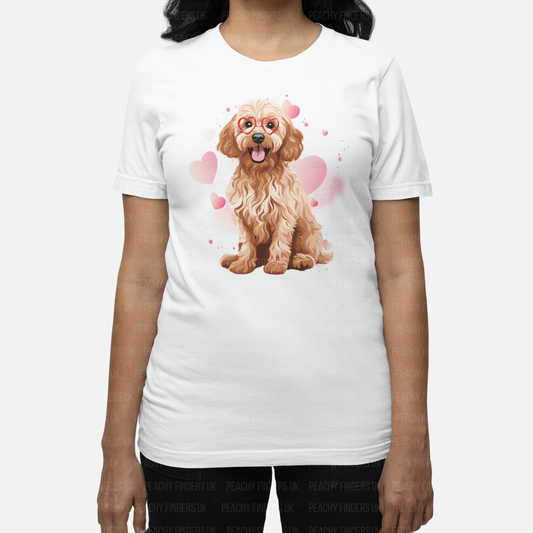 A woman with long dark hair wears a white t-shirt featuring a cartoon Cockapoo dog with red heart-shaped glasses. Pink hearts and subtle pink watercolour-style splashes surround the dog on the shirt.