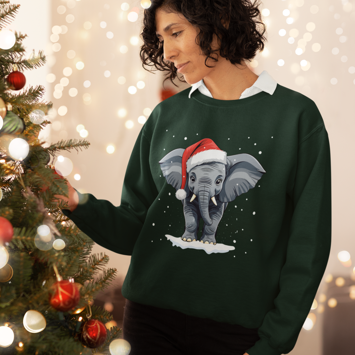 Woman decorating a Christmas tree while wearing a dark green festive sweatshirt featuring a cute baby elephant in a Santa hat with falling snow.