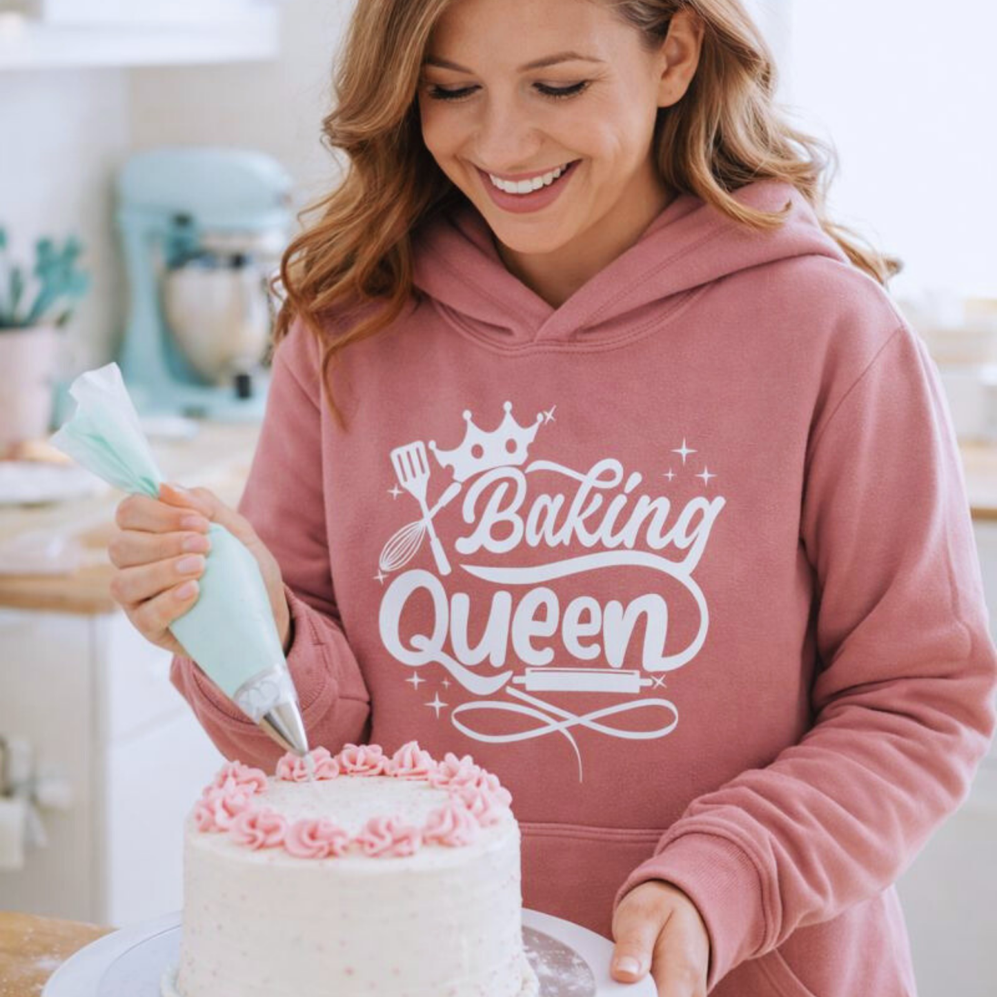 Smiling woman decorating a cake while wearing a dusty pink ‘Baking Queen’ hoodie in a bright kitchen