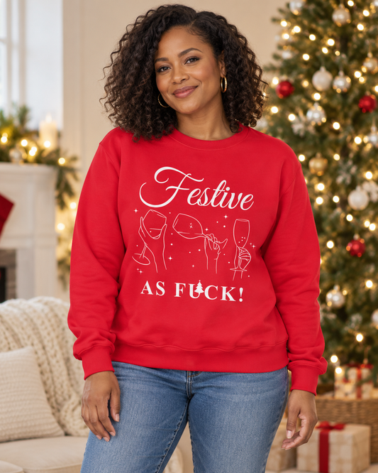 A smiling woman with curly hair wearing a red crewneck sweatshirt with a white graphic that reads 'Festive AS FUCK!' and simple line drawings of wine glasses and champagne flutes. The background shows a softly blurred Christmas tree with warm lights.