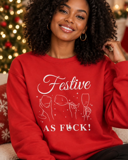 A smiling woman with curly hair wearing a red crewneck sweatshirt with a white graphic that reads 'Festive AS FUCK!' and simple line drawings of wine glasses and champagne flutes. The background shows a softly blurred Christmas tree with warm lights.