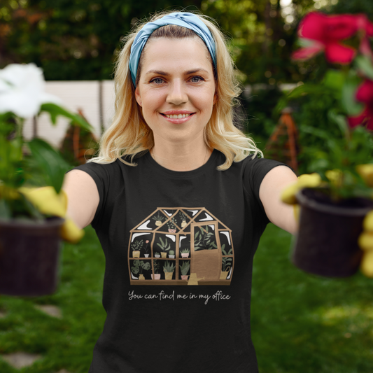 Smiling woman in "You can find me in my office" greenhouse t-shirt holds potted plants. Wearing yellow gloves in the garden.