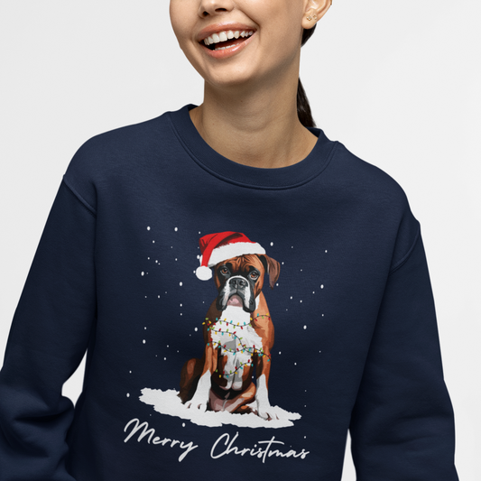 Woman wearing a navy jumper featuring a festive boxer dog wearing a Santa hat, tangled in Christmas lights, sitting in snow with “Merry Christmas” text.