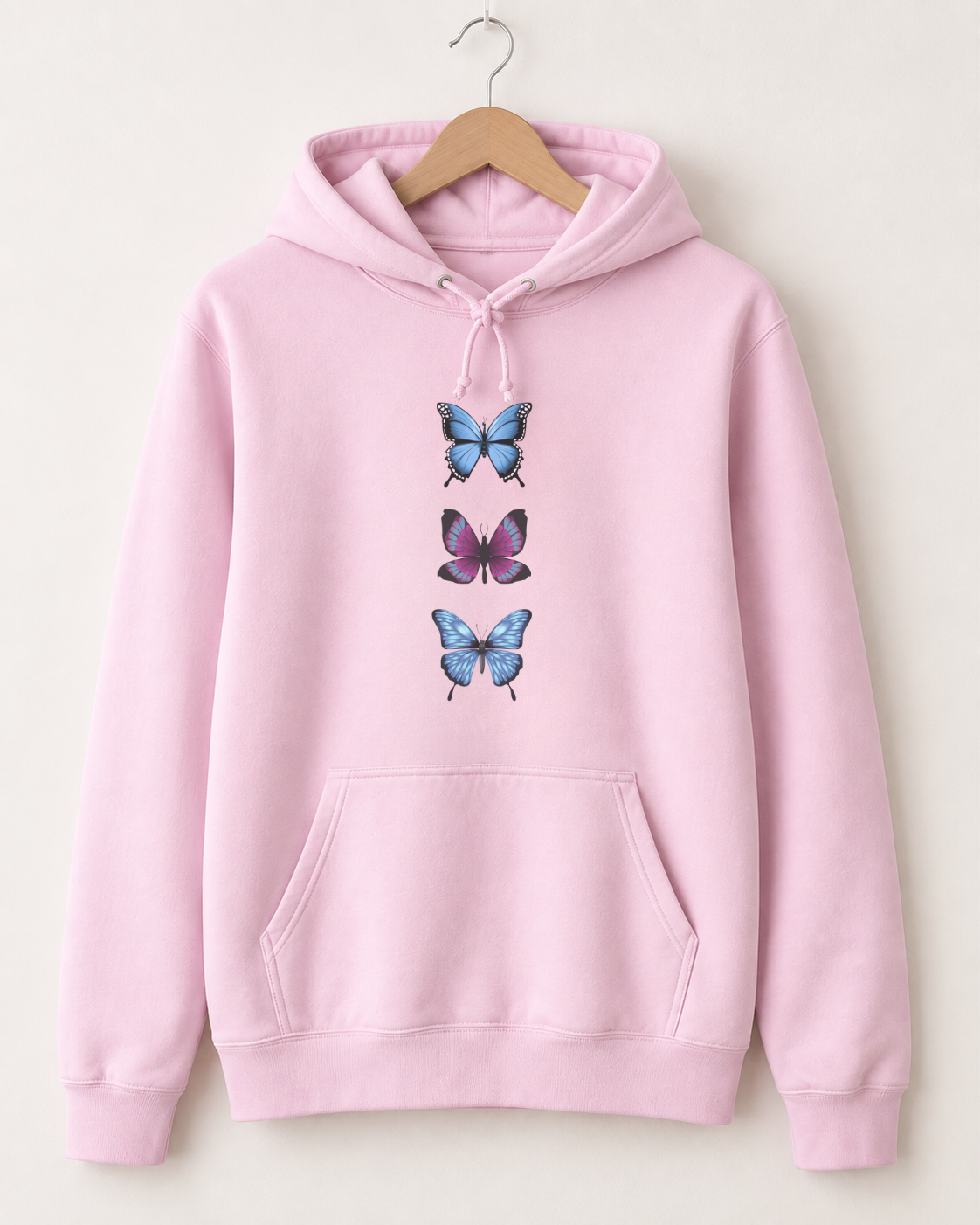 A light pink pullover hoodie hanging on a wooden hanger against a plain off-white background. The hoodie features a graphic print of three stacked butterflies on the center chest, with two blue butterflies and one purple butterfly.