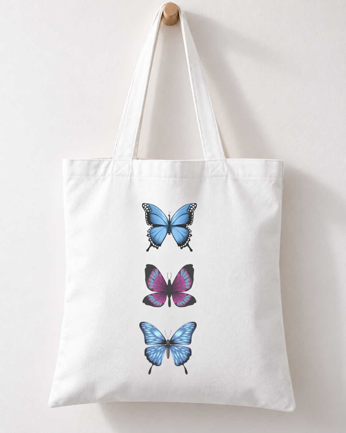 White cotton tote bag with three blue and purple butterflies arranged vertically, hanging by its handles from a wooden wall hook.