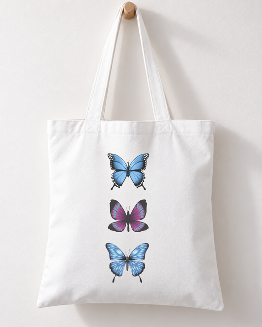 White cotton tote bag with three blue and purple butterflies arranged vertically, hanging by its handles from a wooden wall hook.