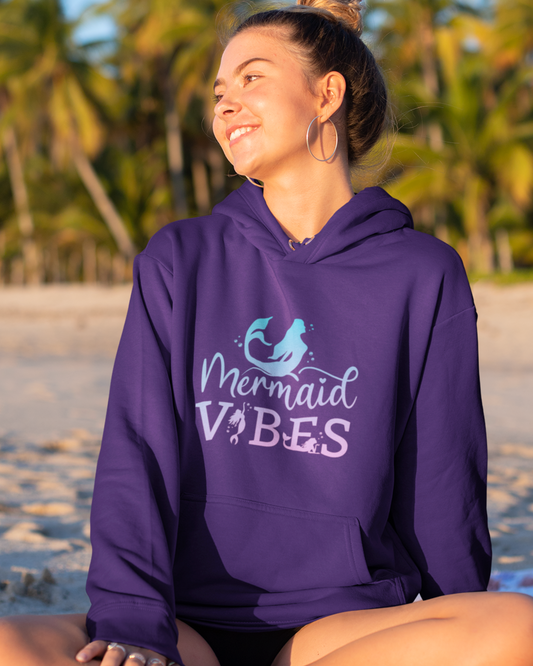 A young woman wearing a purple “Mermaid Vibes” hoodie sitting on a beach at sunset