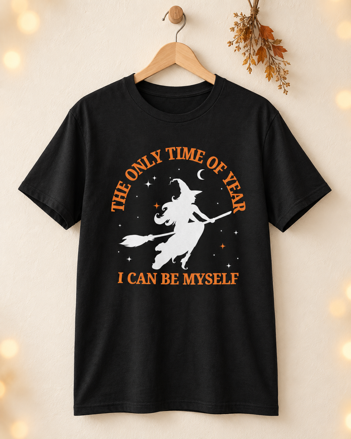 Black flying-witch Halloween T-shirt displayed on a wooden hanger against a warm cream wall.