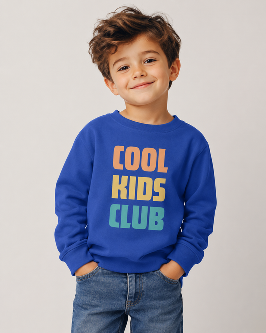 A young boy with brown hair smiling, wearing a royal blue sweatshirt with 'COOL KIDS CLUB' printed in orange, yellow, and teal block letters, paired with blue jeans, against a light grey studio background.