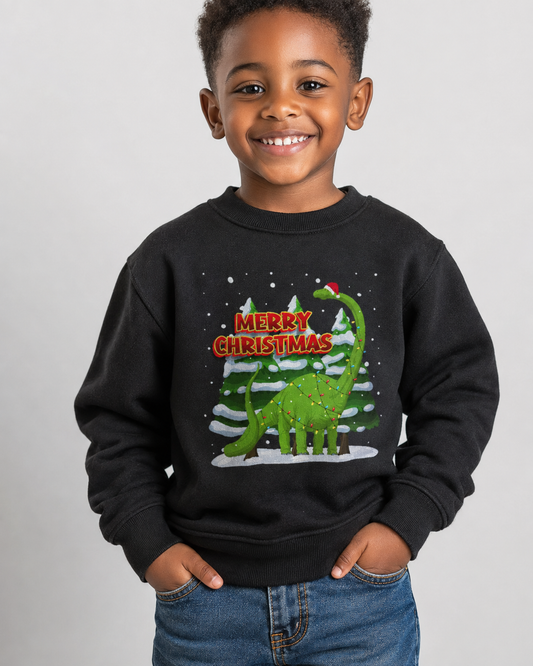 A smiling young boy wearing a black crewneck sweatshirt with a festive Christmas graphic featuring a green dinosaur, snowy pine trees, and 'MERRY CHRISTMAS' text, paired with blue jeans against a light grey background.