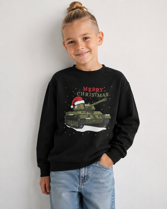 A young boy with a blonde top-knot hairstyle is wearing a black crewneck sweatshirt with a 'Merry Christmas' military tank graphic, paired with light blue jeans, standing against a plain light grey wall.