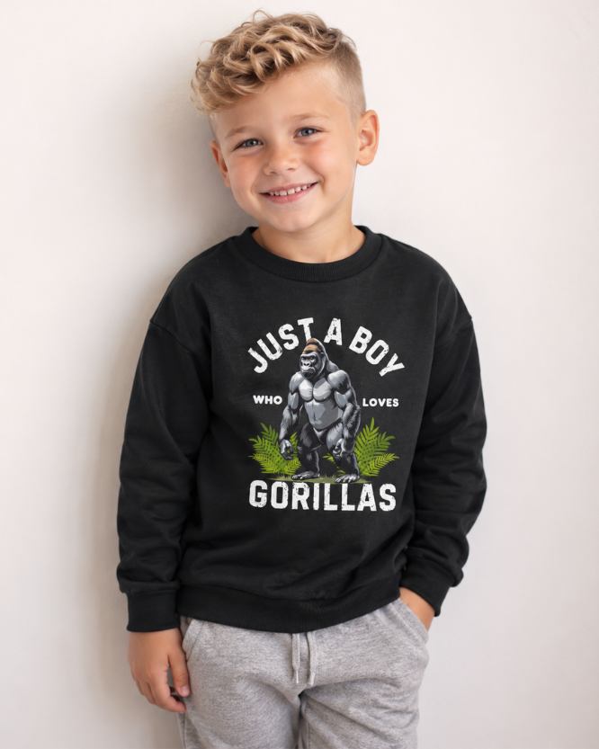 A young boy with blonde hair is smiling and wearing a black crewneck sweatshirt with a graphic of a gorilla and the text 'JUST A BOY WHO LOVES GORILLAS' in white, paired with light gray sweatpants, against a plain off-white wall.