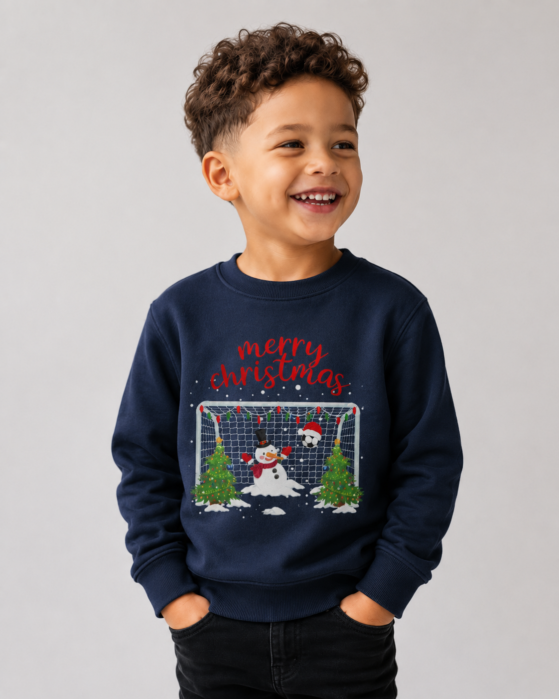 A young boy with curly brown hair smiling, wearing a navy blue crewneck sweatshirt with a festive Christmas graphic. The graphic features the text 'merry christmas' in red script above a scene of a snowman, a soccer ball, a goal net, and two Christmas trees.