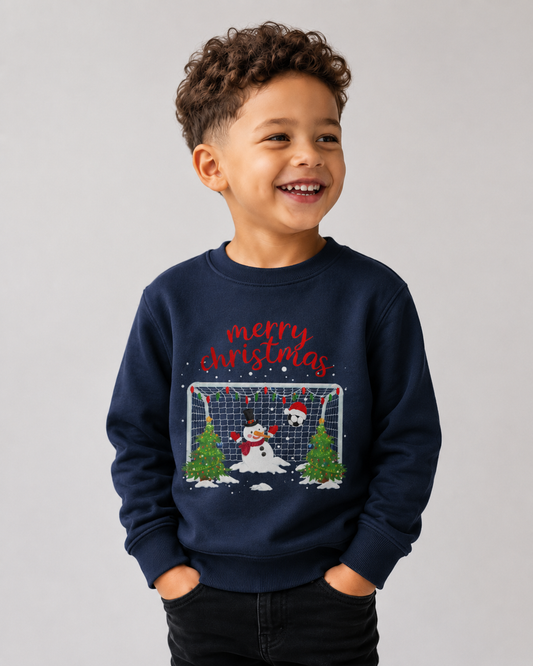 A young boy with curly brown hair smiling, wearing a navy blue crewneck sweatshirt with a festive Christmas graphic. The graphic features the text 'merry christmas' in red script above a scene of a snowman, a soccer ball, a goal net, and two Christmas trees.