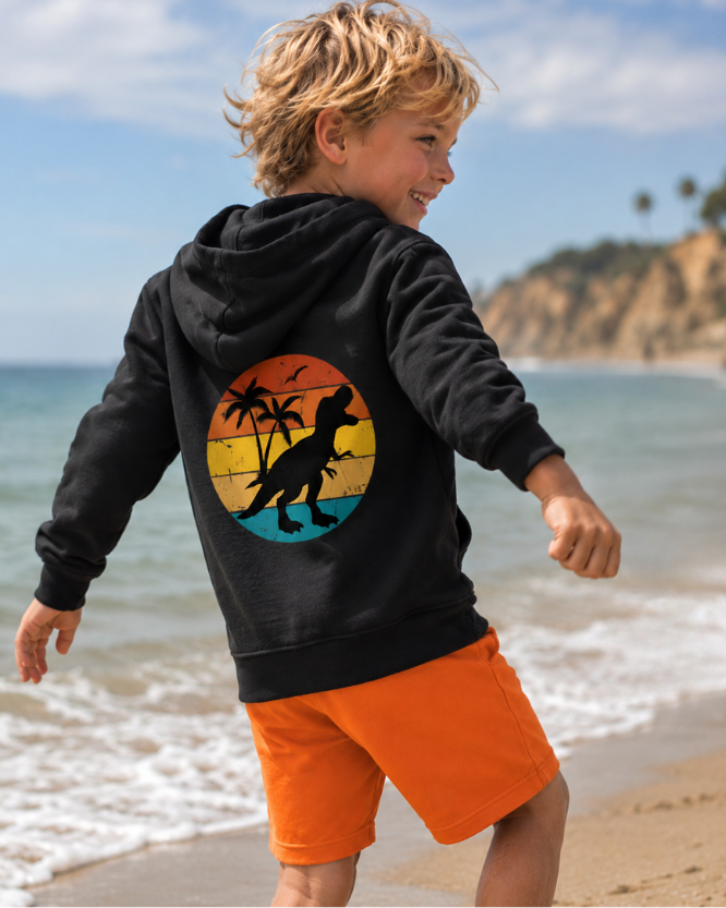 Young boy walking by the sea wearing a black dinosaur zip hoodie with colourful retro T-Rex back design.