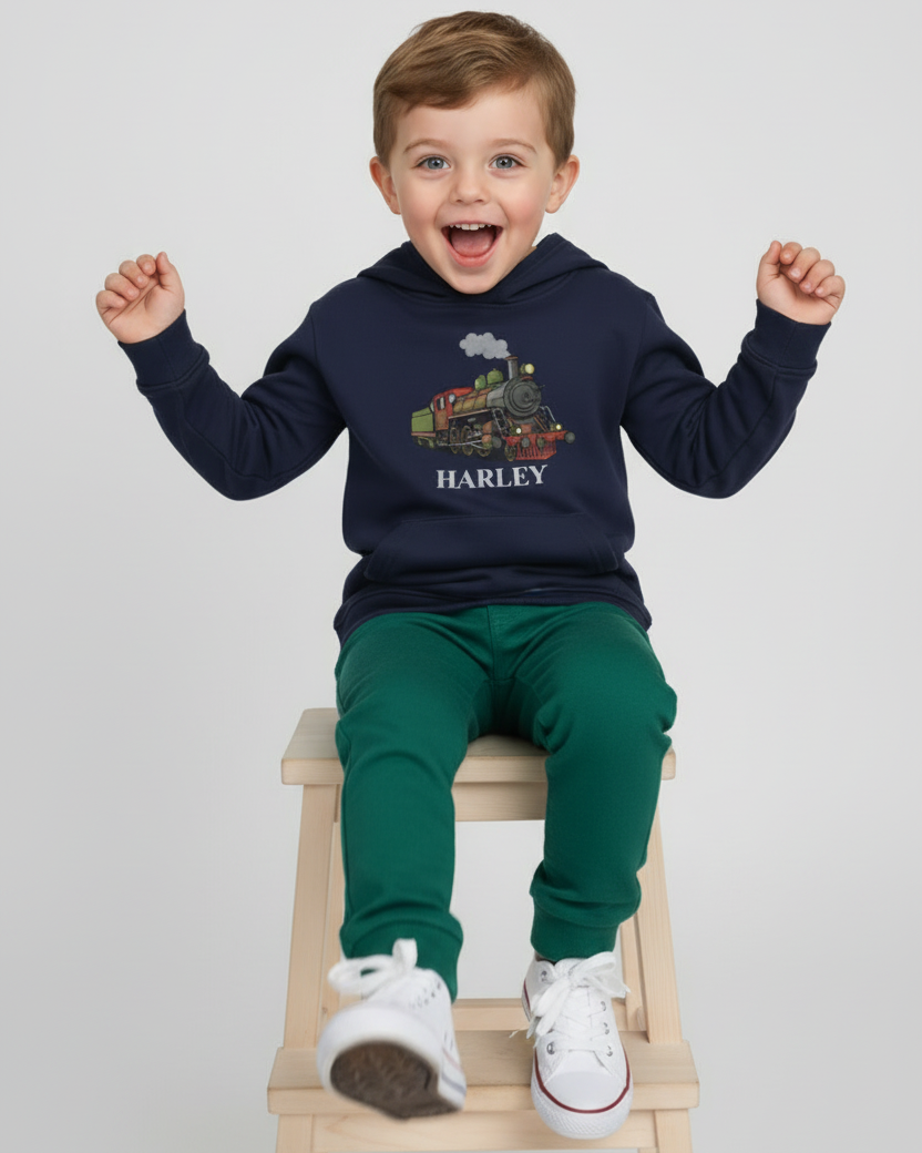 Smiling child model sitting on a stool wearing a navy personalised steam train hoodie, green joggers and white trainers in a bright studio setting.