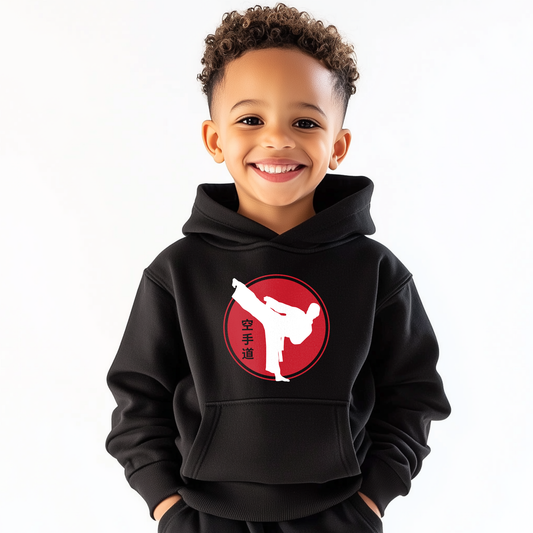 Young boy wearing a kids karate hoodie featuring a white kicking silhouette inside a red circle with Japanese kanji symbols, smiling against a plain white background.