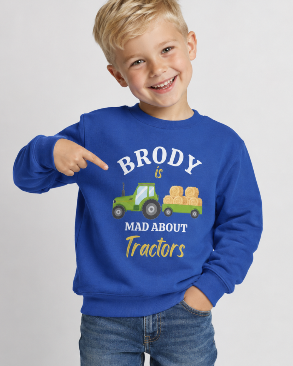 A young boy with blonde hair smiling and pointing at a royal blue sweatshirt. The sweatshirt features a graphic of a green tractor carrying hay bales with the text 'BRODY is MAD ABOUT Tractors' in white and yellow.