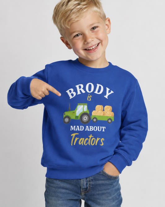 A young boy with blonde hair smiling and pointing at a royal blue sweatshirt. The sweatshirt features a graphic of a green tractor carrying hay bales with the text 'BRODY is MAD ABOUT Tractors' in white and yellow.