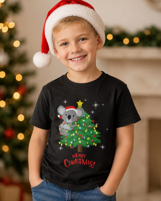 A smiling young boy wearing a red and white Santa hat and a black graphic t-shirt featuring a cute cartoon koala sitting on a decorated Christmas tree with 'Merry Christmas!' written below, posed in front of a softly blurred holiday fireplace background.