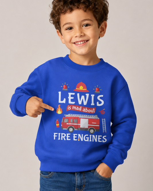 A smiling young boy with curly brown hair wearing a bright blue crewneck sweatshirt with a custom fire engine graphic and the text 'LEWIS is mad about FIRE ENGINES'. He is pointing at the graphic with his right hand and wearing blue jeans, set against a plain light grey studio background.