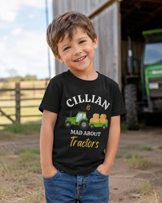 A young boy smiling outdoors in front of a barn and tractor, wearing a black graphic T-shirt that reads 'CILLIAN is MAD ABOUT Tractors' with a green tractor and hay bale illustration.