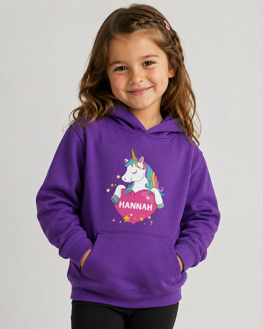 A young girl with brown hair smiles while wearing a purple hoodie featuring a colourful unicorn and heart graphic with the name 'HANNAH' printed inside the heart on the front, set against a plain light grey background.