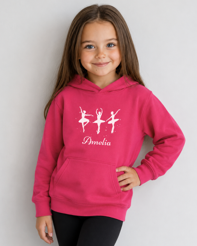 A young girl with long brown hair wearing a hot pink hoodie with white ballet dancer silhouettes and the name 'Amelia' printed on the front, paired with black leggings, posing against a light gray background.