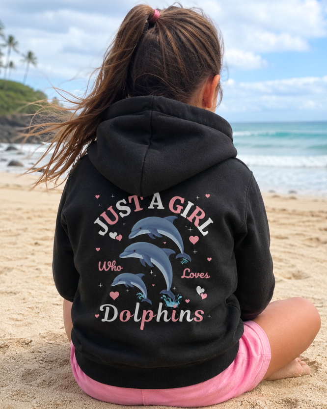 A young girl sitting on a sandy beach facing the ocean, wearing a black zip hoodie from the back which features a graphic of three jumping dolphins and the text 'JUST A GIRL Who Loves Dolphins' with scattered hearts in pink and white, with pink shorts.