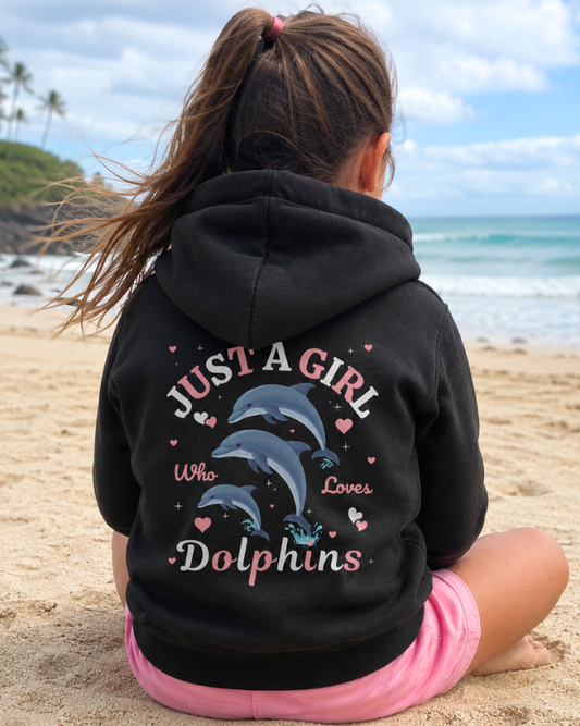 A young girl sitting on a sandy beach facing the ocean, wearing a black zip hoodie from the back which features a graphic of three jumping dolphins and the text 'JUST A GIRL Who Loves Dolphins' with scattered hearts in pink and white, with pink shorts.