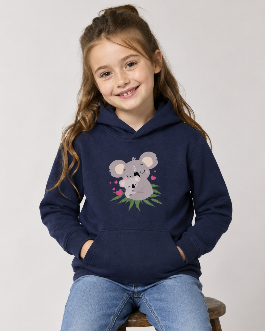 Young girl sitting on a wooden stool in a light studio setting. She wears a navy children’s pullover hoodie and light blue denim jeans featuring a grey mother koala cuddling a baby koala sitting on green leaves, surrounded by small pink hearts.