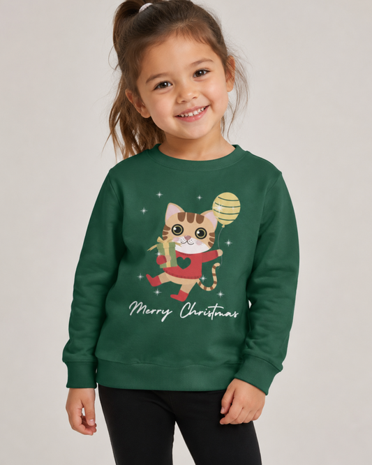 A young girl with a brown ponytail smiles while wearing a dark green crewneck sweatshirt featuring a cartoon cat in a red sweater holding a golden balloon, with 'Merry Christmas' written below. She is wearing black pants against a plain light grey studio background.