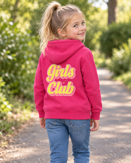 A young girl with blonde hair in a ponytail is shown from the back, wearing a bright pink hoodie with 'Girls Club' printed in yellow and pink bubble letters on the back, paired with blue jeans, standing on a sunlit path surrounded by green trees.