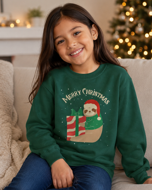 Smiling young girl wearing a dark green kids’ Christmas sweatshirt featuring a sloth in a Santa hat holding a striped present, seated on a sofa beside warm festive lights.