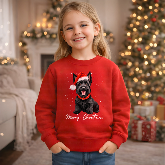 Girl wearing a red kids Christmas jumper with a Scottish Terrier (scottie dog) in a Santa hat and “Merry Christmas” design, festive tree background.