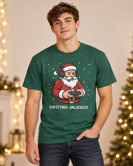 A man wearing a dark green graphic t-shirt with a cartoon illustration of Santa Claus holding a video game controller and the text 'CHRISTMAS UNLOCKED!' below it, set against a warm holiday background with blurred Christmas lights.