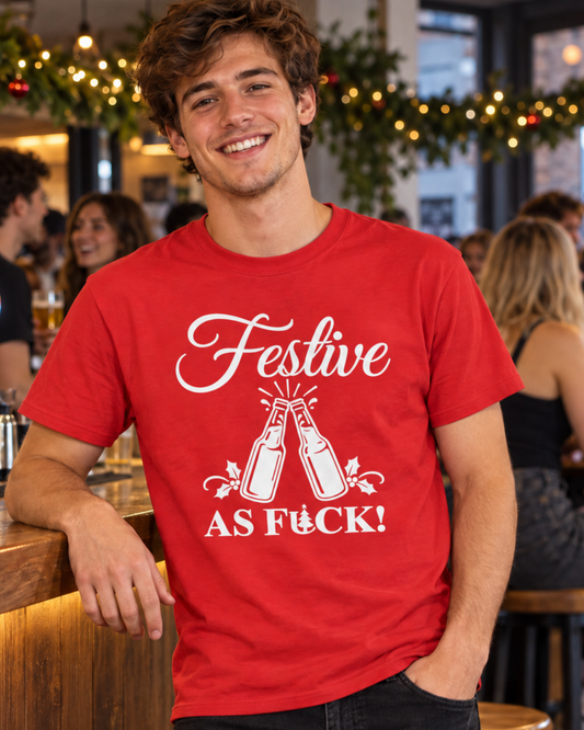 A smiling young man leaning against a bar in a festive social setting, wearing a bright red short-sleeved cotton t-shirt featuring a white graphic of two clinking beer bottles and the text "FESTIVE AS FUCK!".