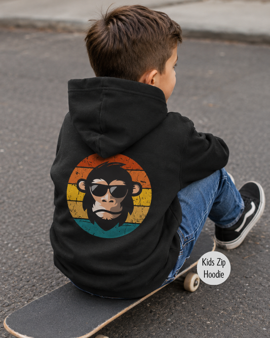 Boy wearing a black kids monkey zip hoodie with a retro chimpanzee back print while sitting on a skateboard outdoors.