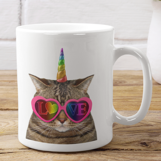 A white ceramic mug with a printed image of a unicorn cat wearing sunglasses and the word 'LOVE' in a rainbow-colored heart.