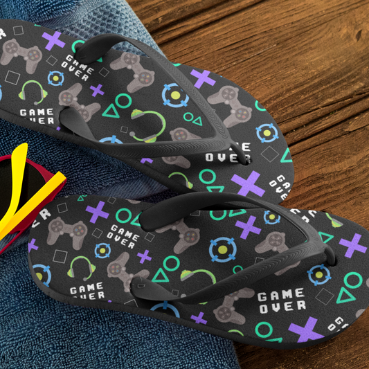 A pair of black kids' flip-flops with a gaming pattern consisting of game icons and text on the straps, displayed on a wooden surface.