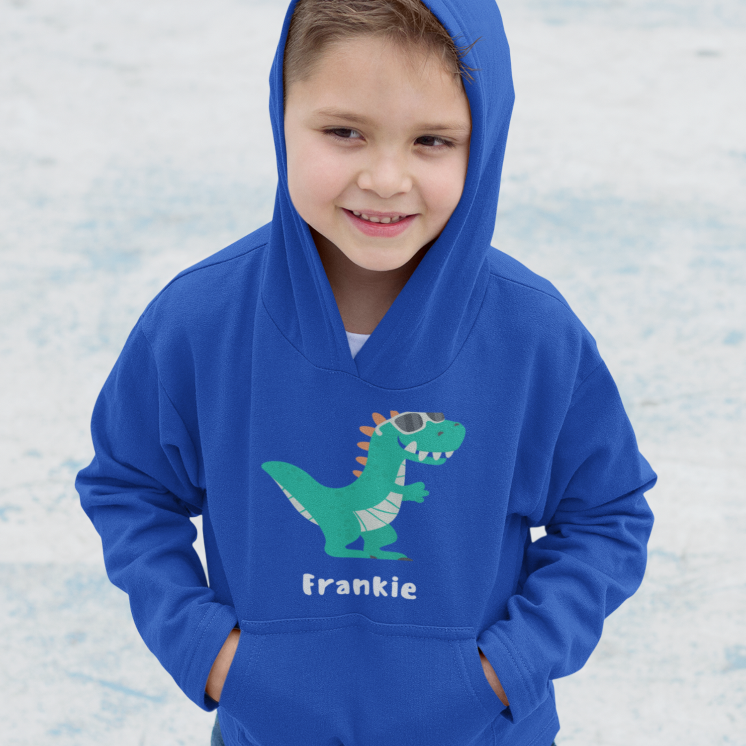 A little boy at the park wearing royal blue pullover hoodie with green dinosaur wearing sunglasses and the personalised name Frankie printed underneath.